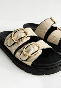 Beige vävda sandaler med tre svarta remmar som har guldbeslag, placerade på en dämpad svart sula med strukturerat mönster.