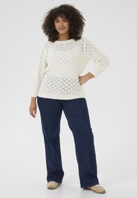 Crème-kleurige, gebreide trui met een ruitpatroon, ronde hals en geribbelde manchetten. Gepaard met donkerblauwe, wijde jeans en beige schoenen.