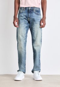 Izbalināti zili džinsi ar taisnām kājām, priekšējām kabatām un vieglu nodilumu; apvienoti ar baltām kedām un gaiši rozā krekliņu.