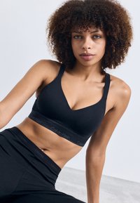 Črni brezžični bralette z v-izrezom, mehkega materiala, širokimi naramnicami in elastičnim trakom s blagovno znamko. V kombinaciji s črnimi hlačami z visokim pasom.