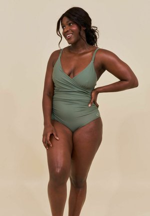 Femeie zâmbitoare cu piele închisă la culoare și păr lung poartă un costum de baie întreg verde măsliniu, stând cu mâna pe șold pe fundal bej.