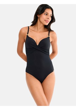 Femme portant un maillot de bain une pièce noir avec un détail en tissu croisé, fines bretelles avec anneaux dorés, debout devant un fond blanc uni.