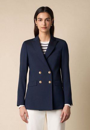 Kobieta ubrana w granatową marynarkę dwurzędową z złotymi guzikami, na białej i granatowej paskowanej bluzce i białych spodniach, stojąca na beżowym tle.