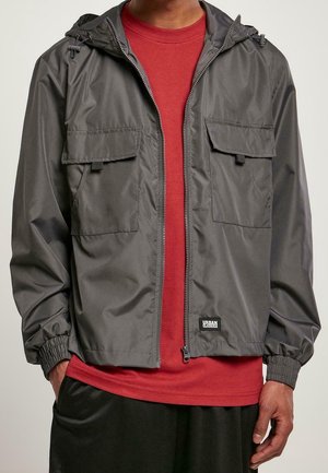 Homme portant une veste à capuche zippée gris foncé sur un t-shirt rouge et un short noir, les mains dans les poches.
