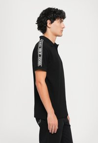 Black polo shirt in cotton with short sleeves, featuring white branded text on the shoulder. Classic collar and relaxed fit design.