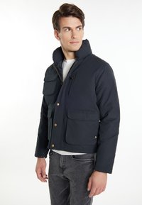 Marineblaue isolierte Jacke mit hohem Kragen, vier Fronttaschen, Reißverschluss und Knopfverschluss sowie einer glatten, matten Oberfläche.