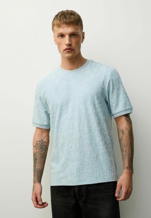 Junger Mann mit kurzen Haaren, der ein hellblaues, strukturiertes Kurzarmhemd, schwarze Hose und geometrische sowie florale Tattoos auf beiden Unterarmen trägt.