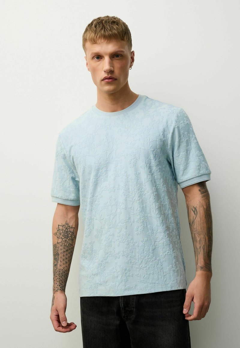 Jeune homme aux cheveux courts portant une chemise à manches courtes texturée bleu clair, un pantalon noir et des tatouages géométriques et floraux sur les deux avant-bras.