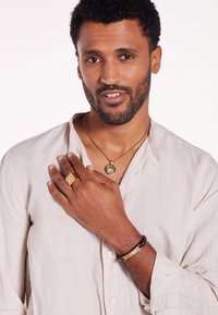 Collier en or avec pendentif, bague en or et bracelet en cuir noir avec des accents dorés. Porté par une personne avec des cheveux courts et bouclés, portant une chemise beige.