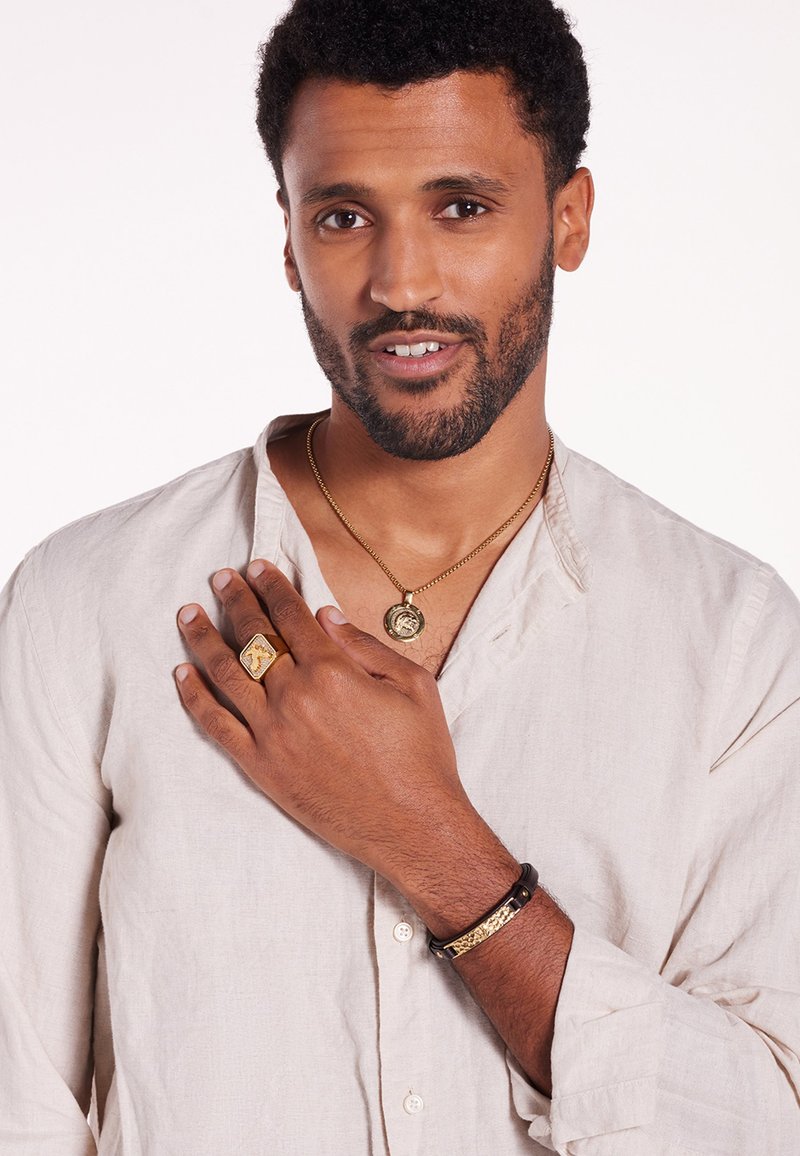 Collier en or avec pendentif, bague en or et bracelet en cuir noir avec des accents dorés. Porté par une personne avec des cheveux courts et bouclés, portant une chemise beige.