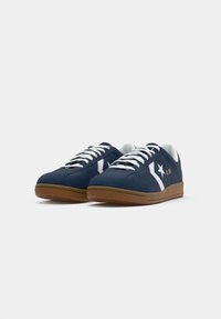 Zapatillas de ante azul marino con detalles y acentos en blanco, que incluyen cordones blancos y una suela de goma marrón. Diseño de perfil bajo.