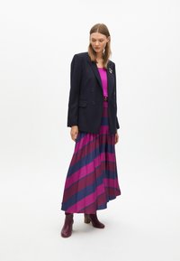 Blazer doppiopetto a righe navy, top fucsia, gonna maxi fluente con audaci righe rosa e viola, stivali alla caviglia bordeaux.