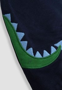 Zils audums ar zaļa un gaiši zilas krāsas izšūtu haizivju zobu dizainu. Gluda tekstūra ar izteiktu krāsu kontrastu.