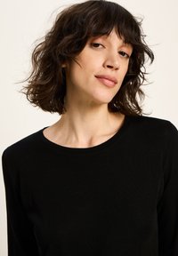 Frau mit kurzem, welligem braunem Haar, die ein schwarzes Langarmshirt trägt und selbstbewusst in die Kamera blickt, vor einem schlichten Hintergrund.