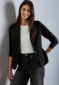 Svart blazer med skräddarsydd passform, två framfickor och silverfärgade knappar, i kombination med en vit topp och mörka jeans. Slät tygstruktur.
