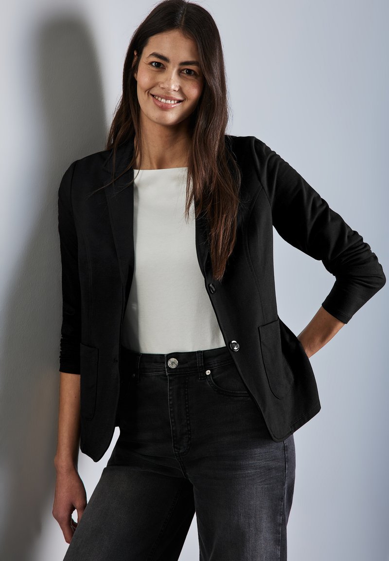 Svart blazer med skräddarsydd passform, två framfickor och silverfärgade knappar, i kombination med en vit topp och mörka jeans. Slät tygstruktur.