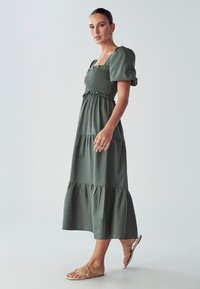Zelena, duga midi haljina s napuhanim rukavima i smockanim korzetom. Ima višeslojni dizajn, meku tkaninu i ležerni siluetu. Nosila se uz sandale s trakama.