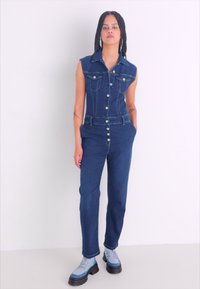Combinaison en denim avec design sans manches, devant à boutons et poches latérales. Tissu bleu foncé avec coutures claires. Mannequin portant des bottes grises et noires.