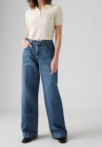 Femme portant un polo crème à manches courtes rentré dans un jean bleu taille haute à jambes larges avec une ceinture en tissu et des chaussures noires.