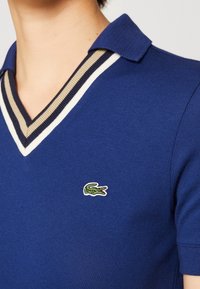Blaues Poloshirt aus strukturiertem Stoff mit V-Ausschnitt, kontrastierendem gestreiften Kragen in Marineblau und Beige sowie kleinem, gesticktem grünen Krokodil-Logo.