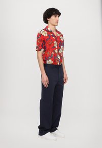 Camisa roja de manga corta con estampado floral y detalles en azul y amarillo, combinada con pantalones navy y zapatillas blancas; ajuste relajado, detalle de botones visible.