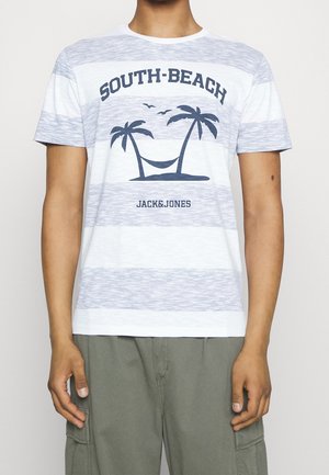Homme portant un t-shirt blanc à rayures bleu clair avec le texte "South Beach" et un motif de palmier avec hamac et oiseaux, associé à un pantalon cargo gris.
