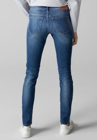 Hubené modré džíny z denimu s mírným vyblednutím, designem na pět kapes a koženou nášivkou na pase. Nosené s bílými teniskami.