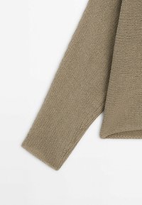 Pull beige en maille avec un tissage lâche, manches longues et ourlets côtelés, présentant un design texturé et une coupe décontractée.