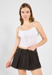 Weißes ärmelloses Oberteil mit Rüschensaum und Lochmuster, kombiniert mit einem dunkelblauen Jeansrock mit Falten und fransigem Saum.