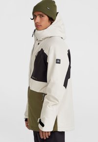 Veste imperméable blanche et vert olive avec des accents noirs, poches zippées et capuche ajustable, fabriquée dans un matériau lisse et durable.
