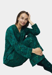 Una joven en pijamas verdes con estampados, sentada con una mano en la cabeza, luciendo relajada contra un fondo liso.