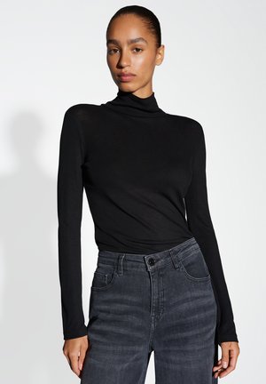 Eine Frau mit zurückgezogenem Haar, die einen eng anliegenden schwarzen Rollkragenpullover und hochgeschnittene dunkelgraue Jeans trägt, vor einem schlichten hellen Hintergrund.