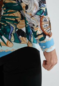 Chemise colorée avec des motifs de papillons et de fleurs, fabriquée en tissu léger avec des poignets bleus et des détails de boutons. Coupe élégante.