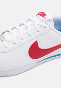 Baltos sportinės bateliai su raudonu Nike swoosh, tekstūruota odinė viršutinė dalis, kontrastingi mėlyni akcentai ir standartinis raištelių uždėjimo dizainas.