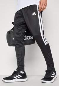 Sorte træningsbukser med hvidt tre-stribe design, lavet af glat stof. Parret med sorte sneakers med hvide detaljer.