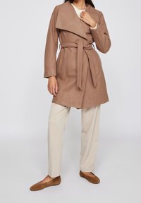Brun bälte omslagsjacka med bred krage, kombinerad med ljusbeige byxor och bruna lägenheter, på en neutral bakgrund. Mjuk textur.