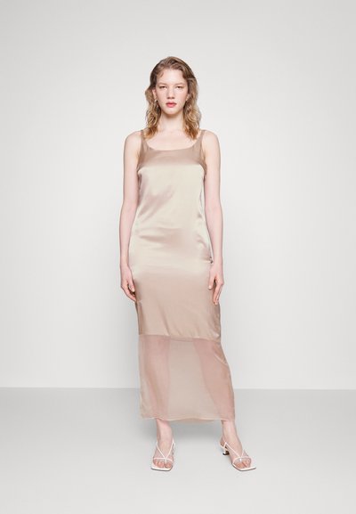 Σατινέ φόρεμα σε ανοιχτό μπεζ χρώμα, με εφαρμοστό τοπ και διαφανές κάτω μέρος. Το μοντέλο φοράει λευκές σανδάλια με τιράντες, στέκοντας απέναντι από έναν κενό φόντο.