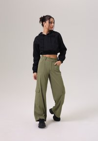 Zwarte cropped hoodie met trekluitjes, gecombineerd met olijfgroene wijde cargobroek met grote zijzakken en een knoopsluiting. Zwarte sneakers.
