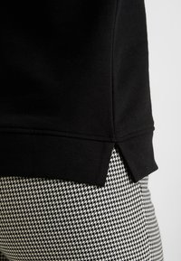 Sudadera negra con dobladillo acanalado y aberturas laterales, combinada con tejido de patrón de pata de gallo en blanco y negro. Textura suave.