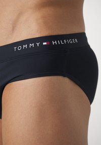 Bañadores de natación en color marino con una cinturilla ancha que presenta el logo "TOMMY HILFIGER" en acentos blancos y rojos. Tejido suave y elástico.