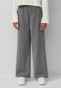 Graue weite Hosen aus strukturiertem Stoff mit einem elastischen Bund. Getragen mit weißen Sneakers vor einem neutralen Hintergrund.