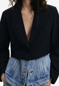 Schwarzer kurz geschnittener Blazer mit tiefem V-Ausschnitt, einreihiger Knopfleiste und strukturierten Schultern, kombiniert mit einem knopfbesetzten Jeansrock mit sichtbaren Nähten.