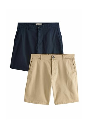 Junger Mann steht an einer schlichten Wand, trägt ein schwarzes Sweatshirt, beige Shorts, weiße Socken und beige Sneaker.