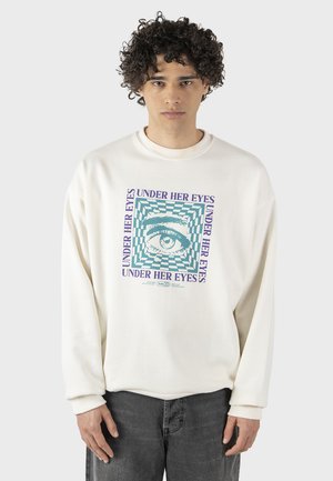 Crème-kleurige sweatshirt met een centraal grafisch ontwerp van een oog in een vierkant patroon, aangevuld met paarse tekst "UNDER HER EYES."