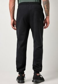 Mand iført sorte joggingbukser og sorte sneakers, set fra ryggen med en lomme på højre side og en synlig tatovering på armen.