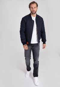 Giacca bomber navy con polsini a coste e zip, abbinata a jeans grigi e sneakers bianche. Presenta un'etichetta tessuta sulla manica della giacca.