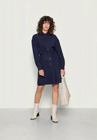 Vestido azul-marinho com botões, de mangas longas, cintura ajustada e comprimento até ao joelho. Usado com botas bégeis até ao tornozelo e segurando uma bolsa leve.