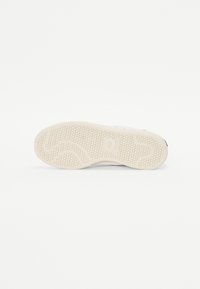 Sola de ténis branca com uma superfície de borracha texturizada. Apresenta um padrão de piso ondulado e um logótipo circular perto do calcanhar.