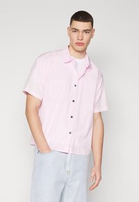Camisa de manga corta rosa hecha de tela ligera, con cuello, bolsillo frontal y botones oscuros, combinada con unos vaqueros de mezclilla azul claro.