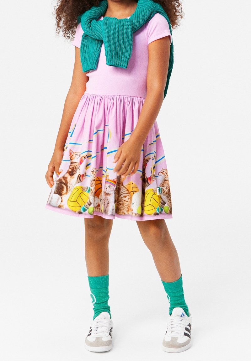 Fille portant une robe rose avec un motif de chat et de ballon de sport, un pull vert noué autour des épaules, des chaussettes vertes et des baskets blanches.